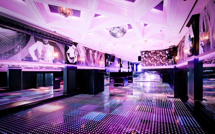 Empty retro-modern nightclub interior with a glowing dotted LED dance floor, mirrored walls, disco ball and pink-purple ambient lighting over plush booths and geometric ceiling.