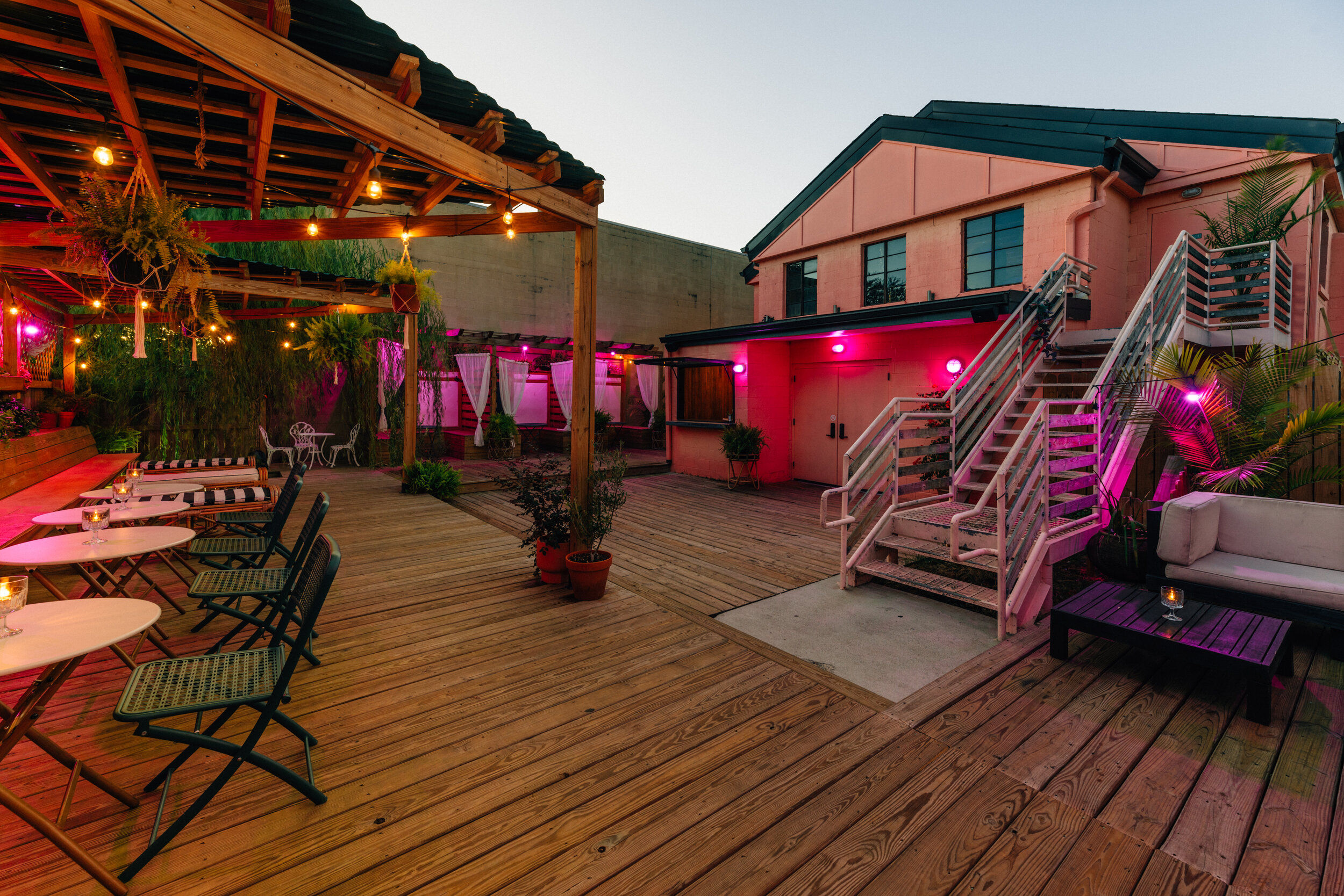 Cozy urban rooftop patio on a wooden deck with pergola and hanging string lights, bistro tables, potted plants, lounge seating and pink-lit staircase at dusk