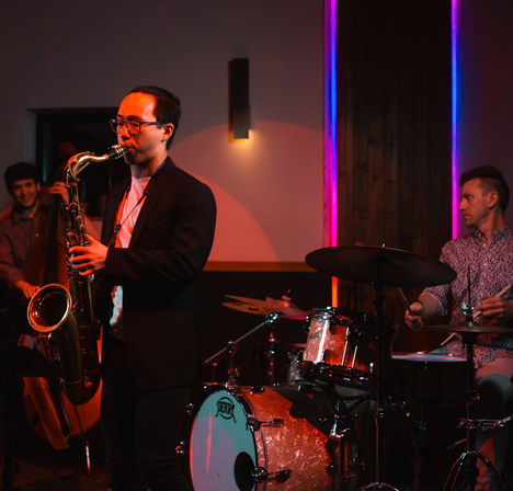 Saxophonist performing with drummer and upright bass in an intimate, dimly lit jazz venue accented by pink and blue neon lighting.
