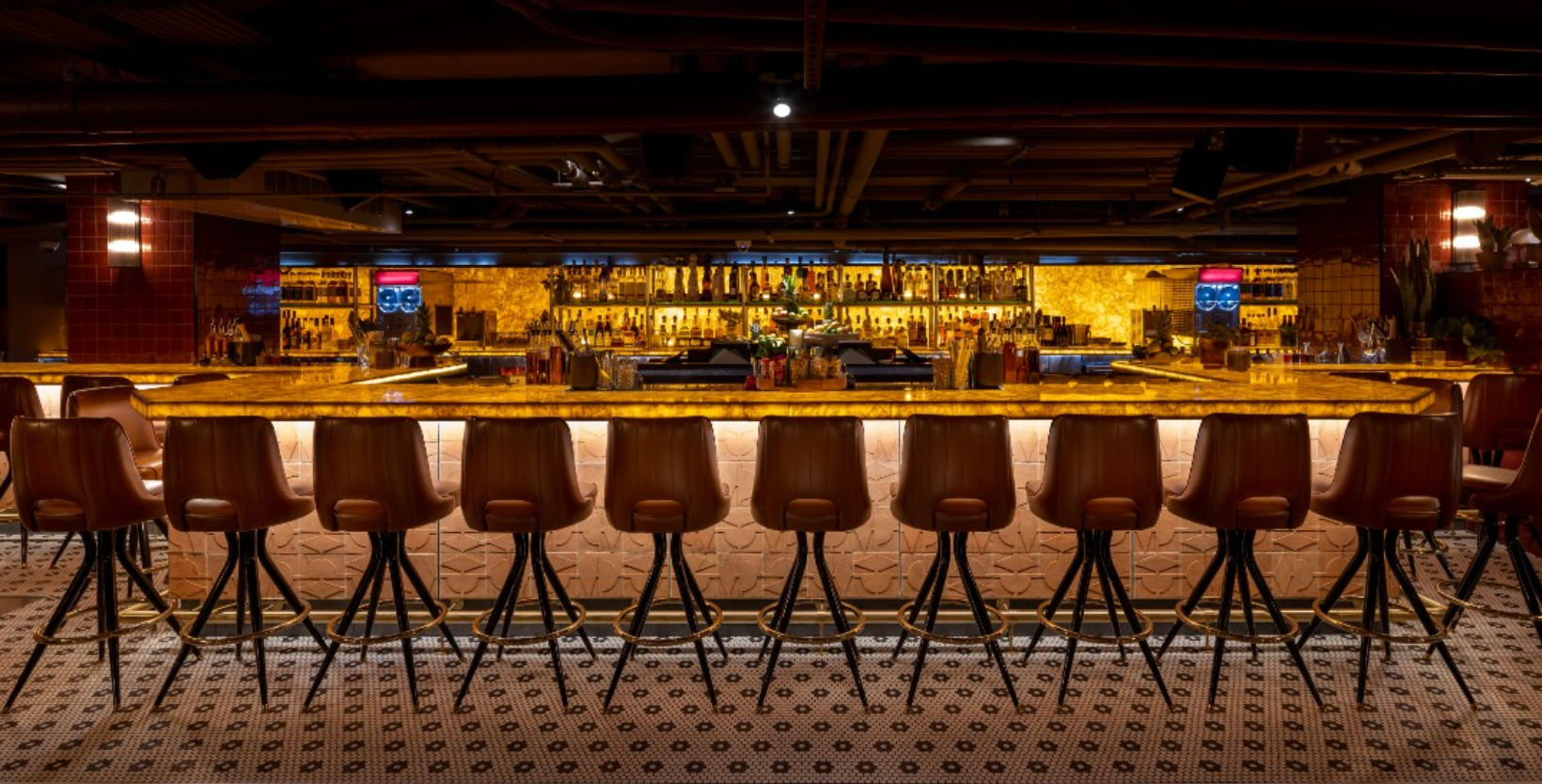 Moody upscale bar interior with a glowing amber backlit counter, a row of brown leather stools, patterned tile floor, and illuminated liquor shelves.