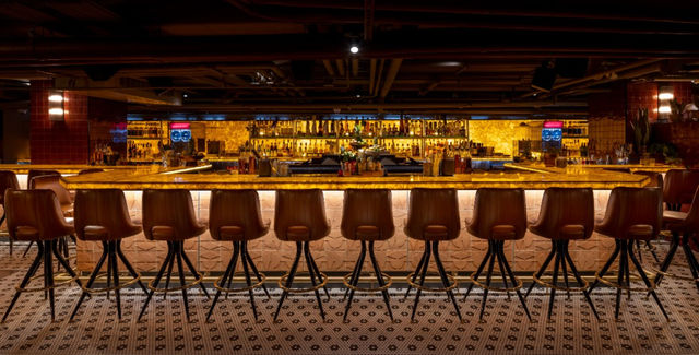 Moody upscale bar interior with a glowing amber backlit counter, a row of brown leather stools, patterned tile floor, and illuminated liquor shelves.