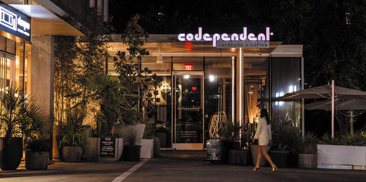 Neon-lit glass-front cocktail and coffee bar entrance at night with potted plants, outdoor umbrellas, and a person in a white jacket walking by on an urban streetscape.