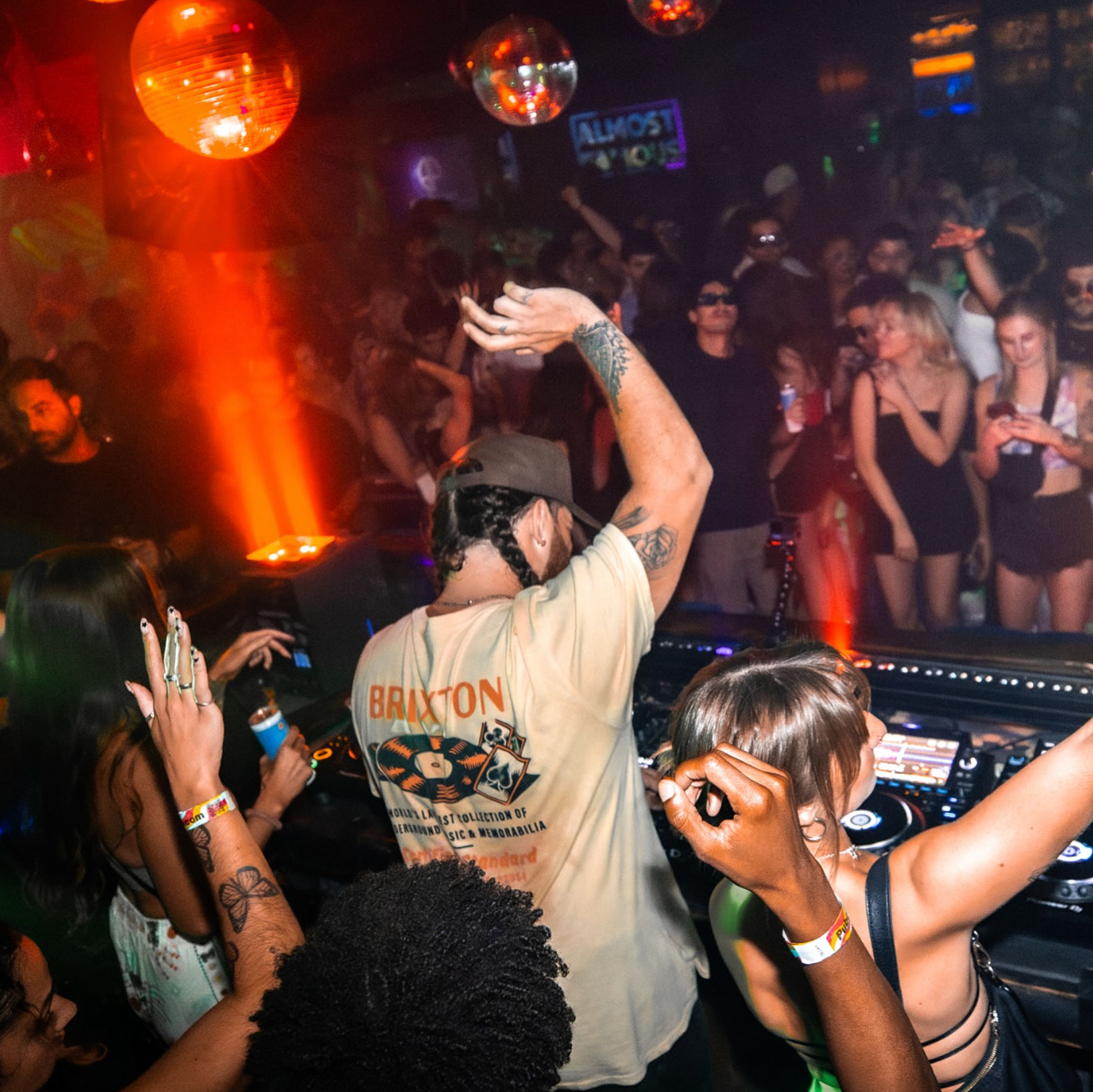 DJ at a crowded nightclub booth raises an arm as partygoers dance under disco balls and warm red lighting — energetic urban nightlife scene