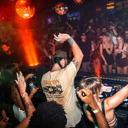 DJ at a crowded nightclub booth raises an arm as partygoers dance under disco balls and warm red lighting — energetic urban nightlife scene