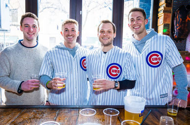 Four friends at a lively bar holding plastic cups of beer, three wearing Chicago Cubs pinstripe jerseys and smiling in front of a sunlit window.