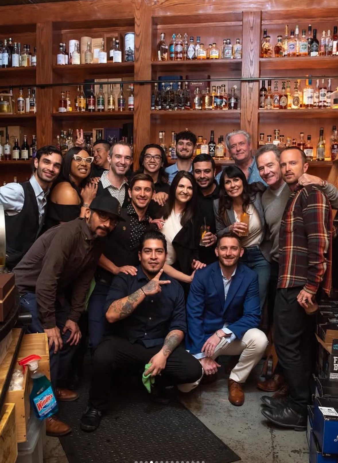 About 16 people smiling and posing in a cozy bar environment in front of wooden shelves packed with whiskey and spirit bottles, some holding cocktails for a casual celebration vibe.