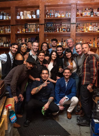 About 16 people smiling and posing in a cozy bar environment in front of wooden shelves packed with whiskey and spirit bottles, some holding cocktails for a casual celebration vibe.