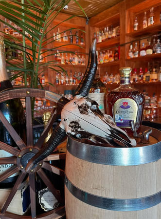 Eye-catching rustic bar display: a horned animal skull perched on a wooden barrel next to whiskey bottles and a ship wheel, warm wooden shelves of spirits blurred in the background.