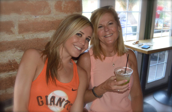 Two smiling women seated by an exposed brick wall and window in a casual cafe, one wearing an orange 'Giants' baseball tank and the other holding a lime cocktail in a plastic cup with a straw.