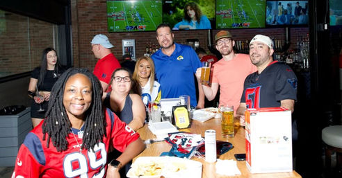 Smiling group at a lively sports bar wearing football jerseys, sharing beers and plates at a wooden table with multiple TVs showing football on a brick wall