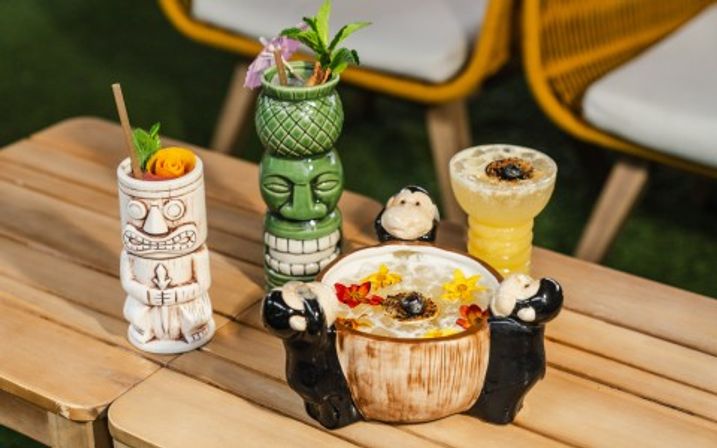 Tropical tiki cocktails in colorful ceramic mugs and a coconut bowl, garnished with edible flowers, mint and citrus, on a wooden outdoor patio table with yellow chairs.