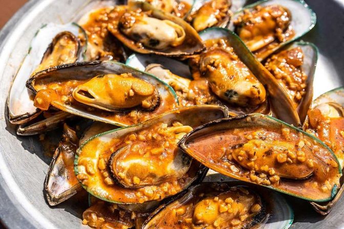 Close-up of a metal platter of mussels in half shells smothered in glossy orange garlic-tomato sauce with minced garlic, savory seafood dish.