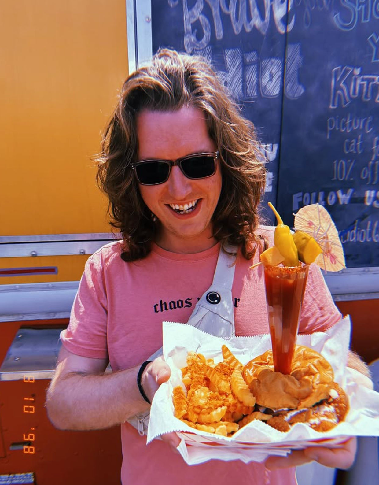 Smiling person in sunglasses and a pink T-shirt holding a food-truck tray with a burger, crinkle-cut fries and fried sides, plus a tall garnished michelada-style drink with peppers and a mini umbrella, chalkboard menu and yellow food truck in the background.