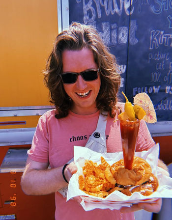 Smiling person in sunglasses and a pink T-shirt holding a food-truck tray with a burger, crinkle-cut fries and fried sides, plus a tall garnished michelada-style drink with peppers and a mini umbrella, chalkboard menu and yellow food truck in the background.