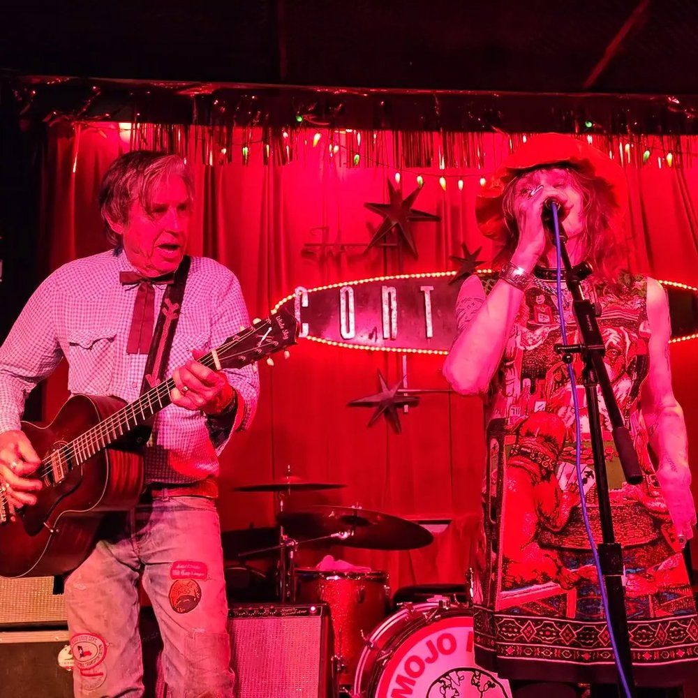 Red-lit intimate club stage with a guitarist strumming an acoustic guitar and a vocalist in a patterned dress and floppy hat singing into a microphone, with drums and a retro illuminated marquee sign behind them.