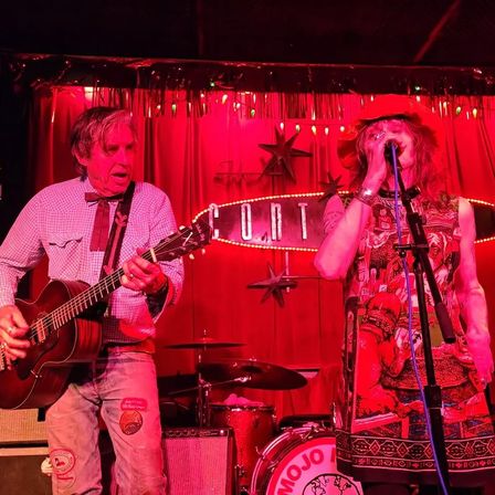 Red-lit intimate club stage with a guitarist strumming an acoustic guitar and a vocalist in a patterned dress and floppy hat singing into a microphone, with drums and a retro illuminated marquee sign behind them.