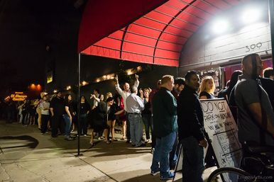 Crowd of people queued outside a busy downtown nightlife venue under a red awning at night, lit by marquee lights and a 'Do Not Park' sign.