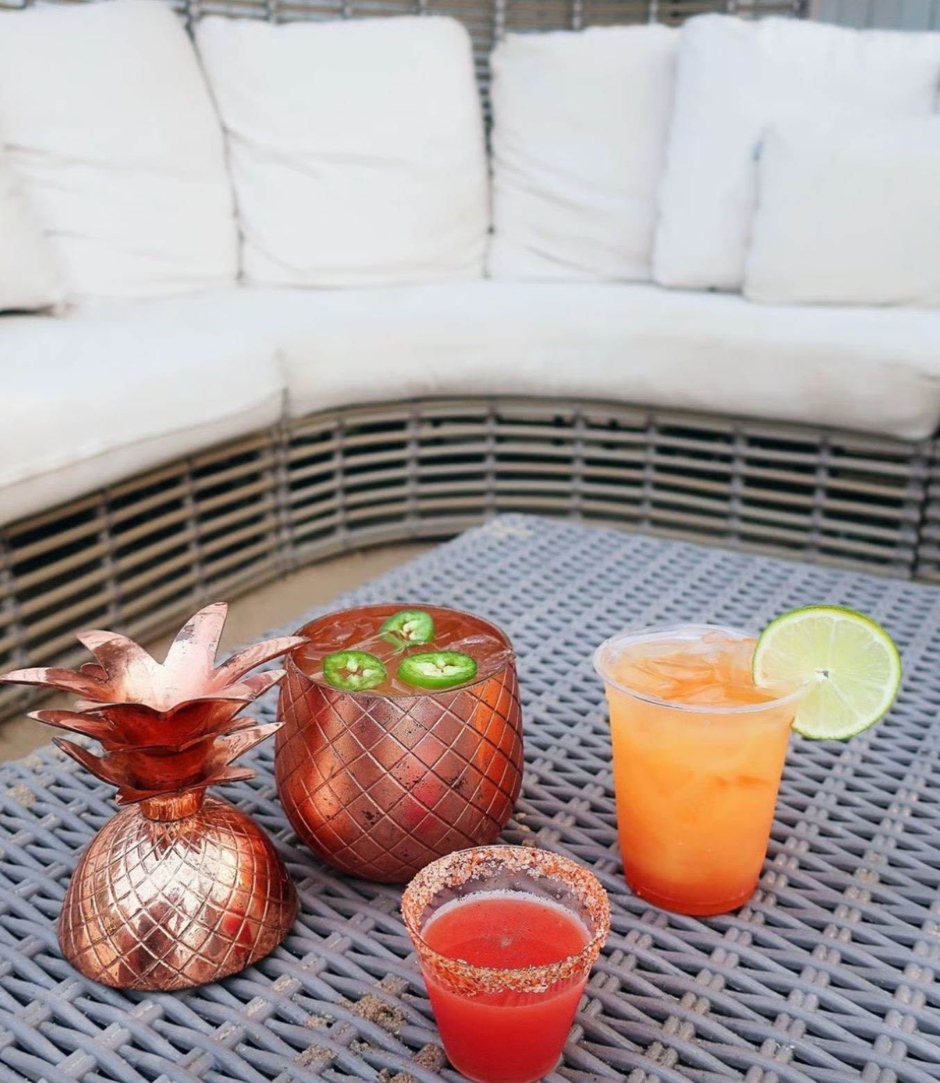 Cozy outdoor patio scene with three colorful cocktails on a gray woven table: a copper pineapple cup topped with jalapeño slices, an orange iced drink with a lime wheel, and a chili-rimmed red shot in front of a white cushioned wicker sectional.