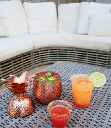 Cozy outdoor patio scene with three colorful cocktails on a gray woven table: a copper pineapple cup topped with jalapeño slices, an orange iced drink with a lime wheel, and a chili-rimmed red shot in front of a white cushioned wicker sectional.