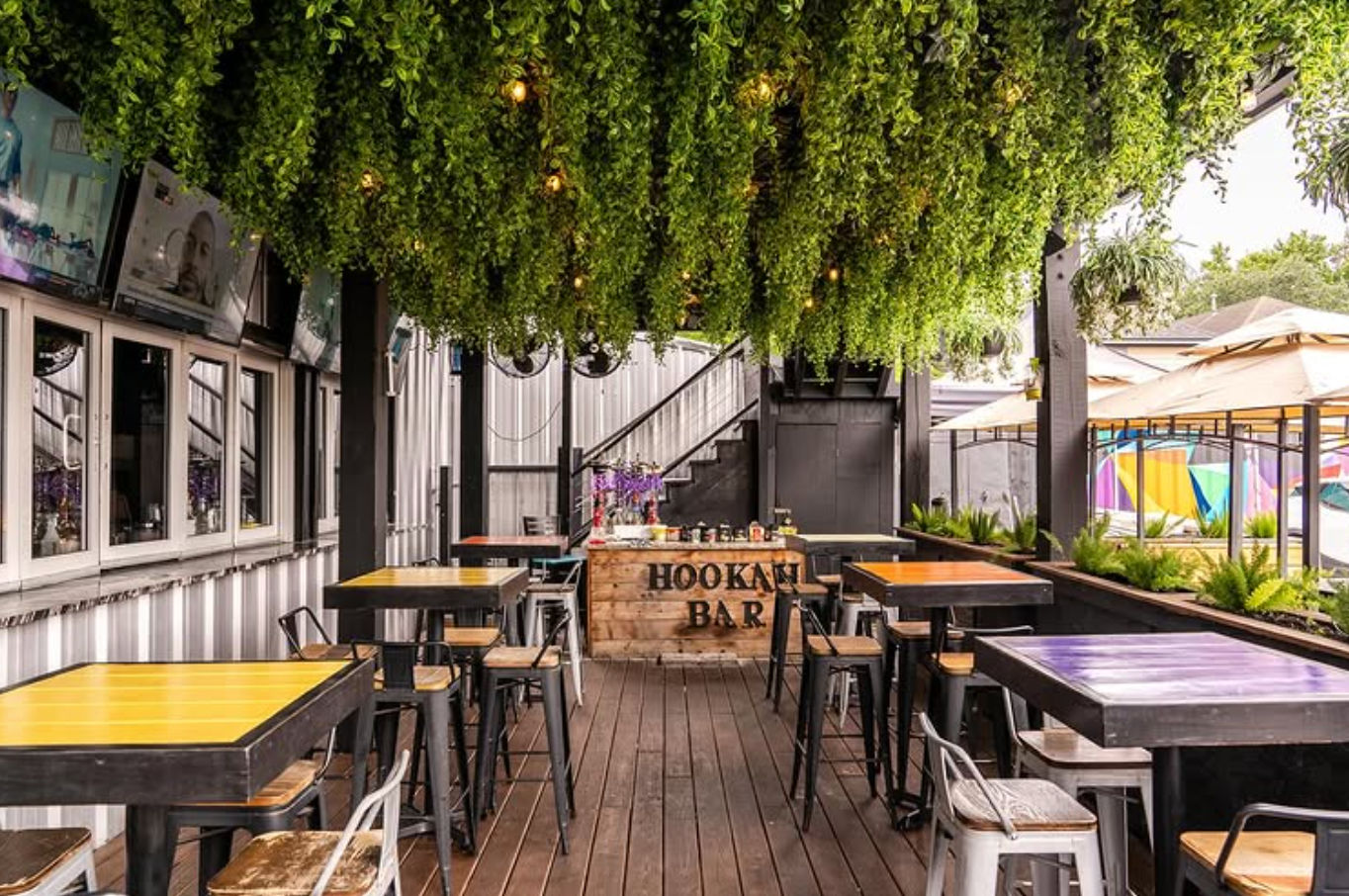 Covered outdoor hookah bar patio in an urban setting with a wooden deck, lush hanging greenery and string lights, colorful high-top tables and metal stools.