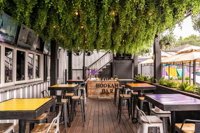 Covered outdoor hookah bar patio in an urban setting with a wooden deck, lush hanging greenery and string lights, colorful high-top tables and metal stools.