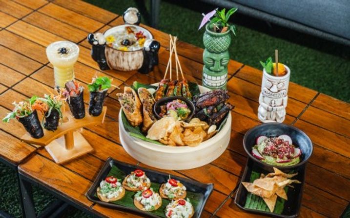 Vibrant tiki-style spread on a wooden outdoor patio table: sushi hand rolls, skewered appetizers, crispy chips, mini tostadas, guacamole bowl and colorful tropical cocktails in tiki mugs.