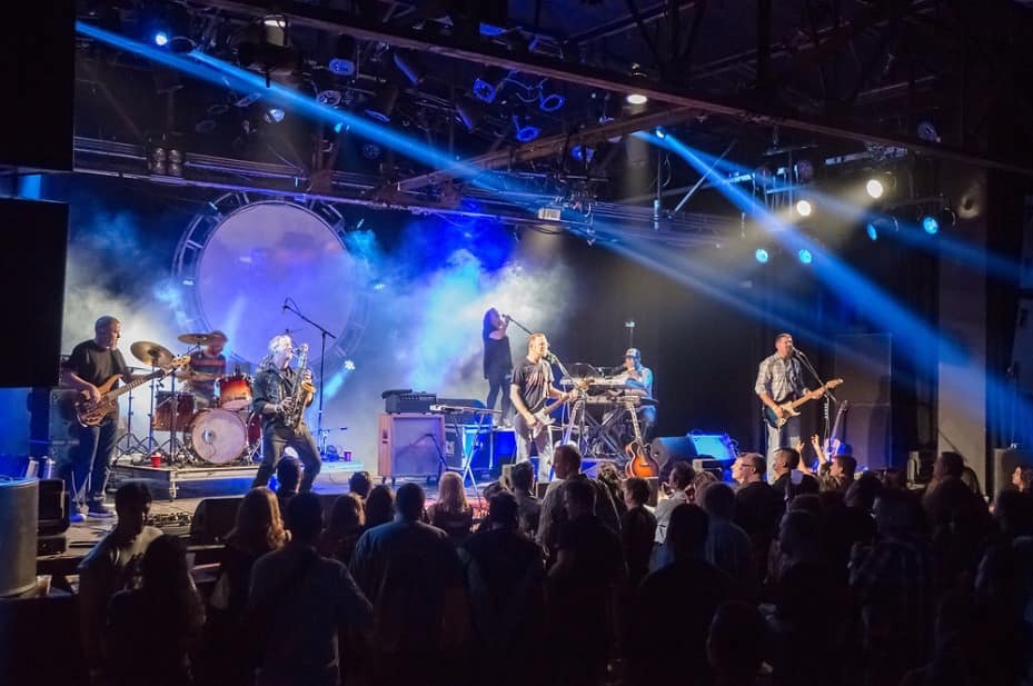 Energetic live band performing at an indoor concert venue with saxophone, guitars, drums and keyboards under blue stage lights and smoke, lively crowd in the foreground