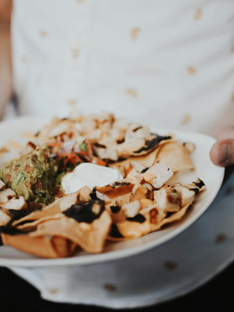 Close-up of Mexican-style loaded nachos on a white plate — crispy tortilla chips topped with melted cheese, grilled chicken, black beans, pico de gallo, guacamole and sour cream, hand holding the plate for casual dining