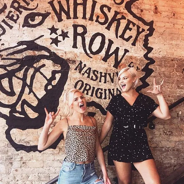 Two smiling young women striking rock-on poses in front of a painted whiskey-themed brick wall mural with a vintage Nashville vibe.