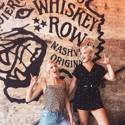 Two smiling young women striking rock-on poses in front of a painted whiskey-themed brick wall mural with a vintage Nashville vibe.