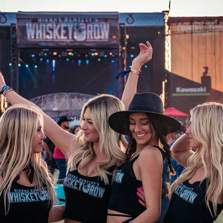 Four friends in matching festival tank tops dancing and smiling at an outdoor country-music concert stage at sunset, one wearing a wide-brim hat