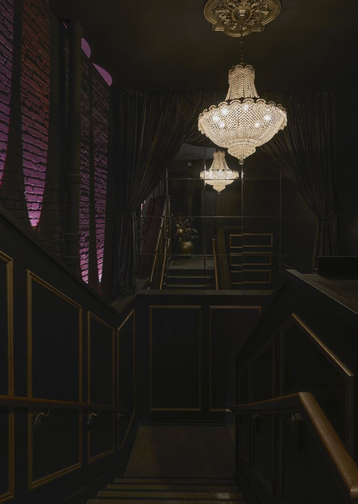Moody, upscale interior stairway with a sparkling crystal chandelier, velvet curtains, mirrored wall and purple-lit exposed brick