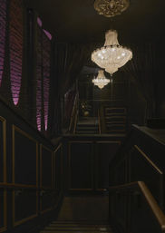 Moody, upscale interior stairway with a sparkling crystal chandelier, velvet curtains, mirrored wall and purple-lit exposed brick