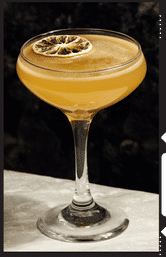 Sunset-hued craft cocktail in a coupe glass topped with a dried citrus wheel, resting on a marble bar countertop against a dark backdrop.