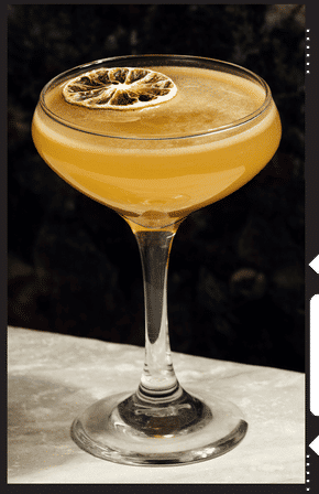 Sunset-hued craft cocktail in a coupe glass topped with a dried citrus wheel, resting on a marble bar countertop against a dark backdrop.