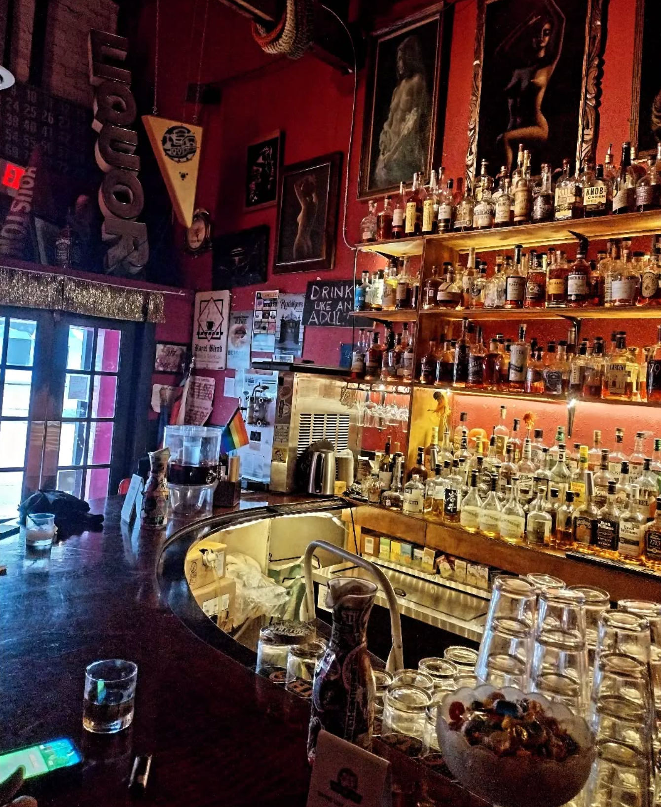 Dimly lit urban bar interior with a curved wooden counter, stacked glasses and bowl of candy, backlit shelves of whiskey and liquor bottles, framed artwork and a small rainbow flag.