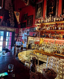 Dimly lit urban bar interior with a curved wooden counter, stacked glasses and bowl of candy, backlit shelves of whiskey and liquor bottles, framed artwork and a small rainbow flag.