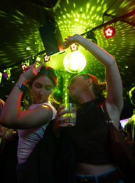 Two women dancing under a green-lit disco ball on a lively nightclub dance floor, colorful spotlights casting dotted patterns while one holds a cocktail glass.