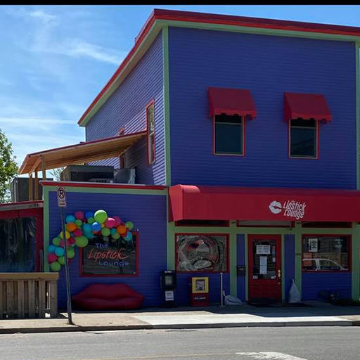 Colorful purple two-story storefront with red awnings and entrance, multicolored balloon arch and lip-shaped bench on a sunny city street.