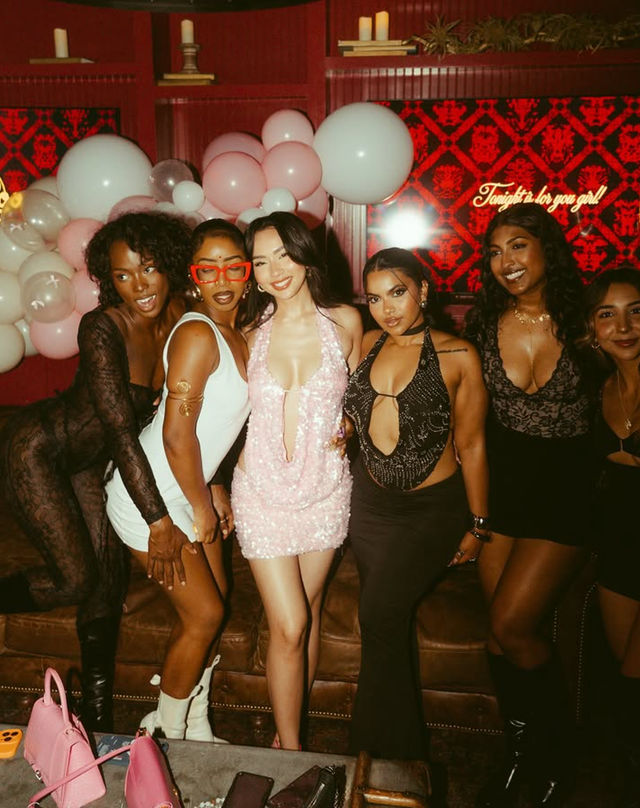 Six friends posing at a pink-themed night out in a dim lounge — center in a sequined pink mini dress, others in stylish club outfits, surrounded by white and pink balloons and a leather sofa.