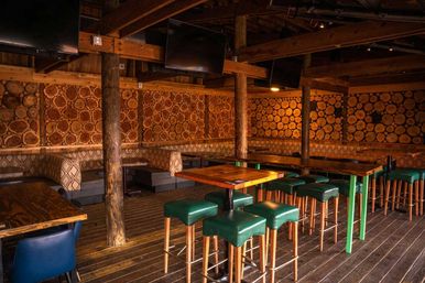 Cozy rustic bar interior with exposed wood beams, log-slice accent wall, warm amber lighting, high-top wooden tables and green upholstered bar stools
