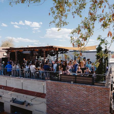 Bustling urban rooftop patio on a sunny day — crowd mingling at a shaded pergola bar atop a brick building with string lights and leafy trees