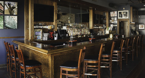Wood-paneled neighborhood bar interior with a polished wooden counter, a row of high-back stools, and a mirrored backbar stocked with liquor bottles and taps.