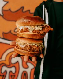 Close-up of a hand holding two stacked crispy fried chicken sandwiches with creamy coleslaw on toasted brioche buns against a vibrant orange street-art mural backdrop