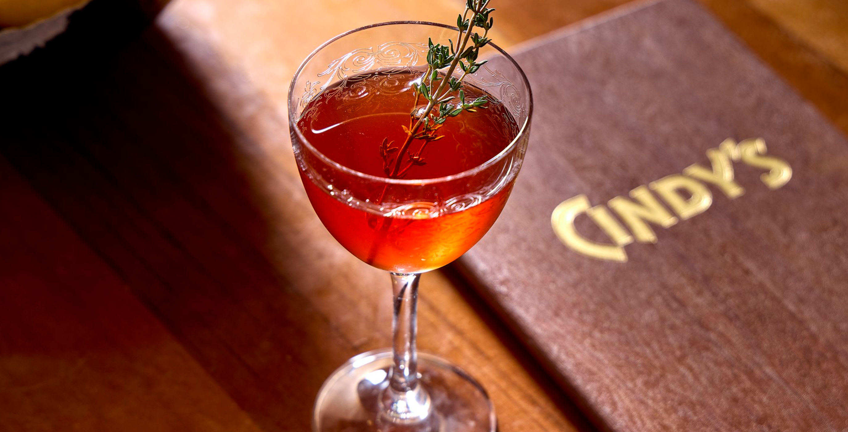 Amber cocktail in an etched coupe glass garnished with a thyme sprig, sitting on a wooden bar table next to a brown menu