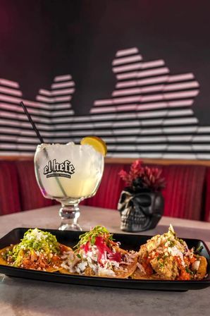 Three colorful street-style tacos topped with shredded meat, pickled onions, crema and fresh herbs on a black plate, paired with a large citrus margarita in a goblet on a table in a dim Mexican restaurant booth with skull décor and striped wall