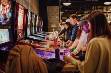 Adults playing a row of vintage pinball machines in a dimly lit arcade bar, beers on the counter and a lively nighttime vibe.