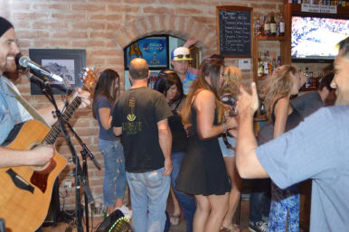 Live music night in a brick‑walled bar — acoustic guitarist singing as a lively crowd chats and dances near the bar with liquor shelves and a TV sports game