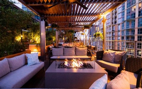 Cozy urban rooftop lounge at dusk with cushioned sofas, central gas fire pit, pergola canopy, hanging plants and high-rise city views — inviting outdoor seating for evenings.