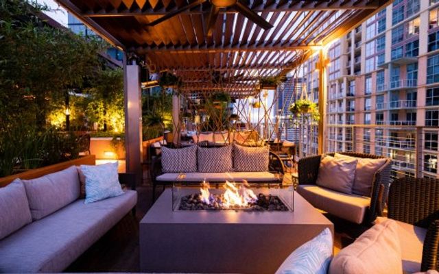 Cozy urban rooftop lounge at dusk with cushioned sofas, central gas fire pit, pergola canopy, hanging plants and high-rise city views — inviting outdoor seating for evenings.